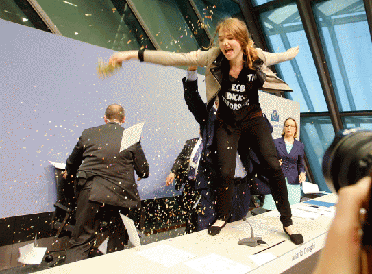 Një aktiviste qëndron mbi tavolinën e podiumit duke i hedhur letra presidentit  të BQE Mario Draghi gjatë një konference të Bankës Qendrore Europiane në Frankfurt, Gjermani. 15 prill 2015. (AP Photo/Michael Probst)