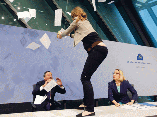 Një aktiviste  qëndron mbi tavolinën e podiumit duke i hedhur letra presidentit  të BQE Mario Draghi , majtas, teksa Christine Graeff, Drejtorja e Përgjithshme e Komunikimeve, sheh  gjatë një konference të Bankës Qendrore Europiane në Frankfurt, Gjermani. 15 prill 2015. (AP Photo/Michael Probst)