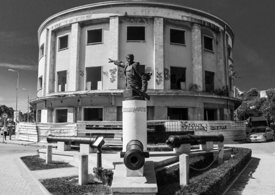 Foto ese: "Shqiptarët e kanë shembur me top vendin e tyre!" "Ish Hotel Vollga në Durrës, një ndërtim i periudhës fashiste, që më vonë u ble nga një tregtar shqiptar para se të shtetëzohej nga regjimi komunist dhe që sot gjendet e rrënuar si pasojë e mosmarrëveshjeve mbi pronësinë. Një investigim i BIRN më herët këtë vit zbuloi prapaskenat e kësaj mosmarrëveshjeje. (Link: http://www.reporter.al/kontrate-me-shullazin-si-u-punesua-i-akuzuari-famekeq-per-konfliktet-e-vollges/) Përballë saj gjendet një bust i stilit soc realist dedikuar Mujo Ulqinakut, ushtarak i Zogut që ra dëshmor në mbrojtje të Durrësit nga pushtimi fashist. Vollga është ndoshta një nga shembujt më tipik se si pasuria e përbashkët apo private kanë përfunduar të rrënuara." Foto Kortezi: Xhodi Hysa