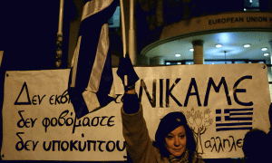 Një grua po valëvit një flamur grek para një banderole ku shkruhet në greqisht “Nuk pranojmë shantazh, nuk kemi frikë, nuk do të mposhtemi, ne fitojmë,” gjatë një proteste kundër BE-së në Nikozia të Qipros më 11 shkurt 2015. Athina dhe Brukseli nuk kanë arritur marrëveshje për programin e shpëtimit nga falimenti i cili skadon më 28 shkurt. (AP Photo/Petros Karadjias)