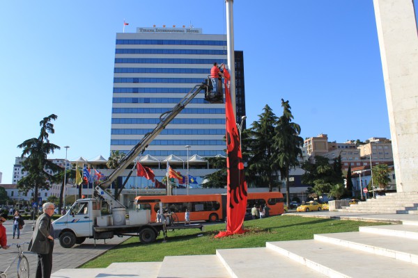 Dy punonjës ngrenë flamurin shqiptar në qendër të Tiranës, teksa një i tretë majtas mbikëqyr punën dhe ndalon fotografimin. Foto: Ivana Dervishi | BIRN.