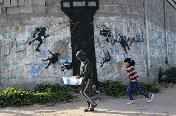 Fëmijë palestinezë po ecin pranë një muraleje ku paraqiten fëmijë duke përdorur një kullë vrojtimi të ushtrisë së Izraelit si një karusel. Grafiti besohet se është pikturuar nga artisti britanik i rrugës Bansky në rrugën kryesore në Beit Lahija në veri të Rripit të Gazës. Një film i shkurtër i publikuar në faqen e tij të internetit, artisti duket se ka hyrë në Gaza përmes një tuneli nëntokësor nga Egjipti. (AP Photo/Adel Hana)