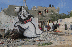 Një grua palestineze po punon bashkë me fëmijët e saj pranë një muraleje që paraqet një kotele të gjallë, e cila prezumohet se është pikturuar nga artisti britanik i rrugës Banksy. Piktura gjendet në një mur të një shtëpie të shkatërruar në luftën mes Izraelit dhe Hamasit verën e kaluar në Beit Hanoun në veri të Rripit të Gazës më 27 shkurt 2015. (AP Photo/Adel Hana)