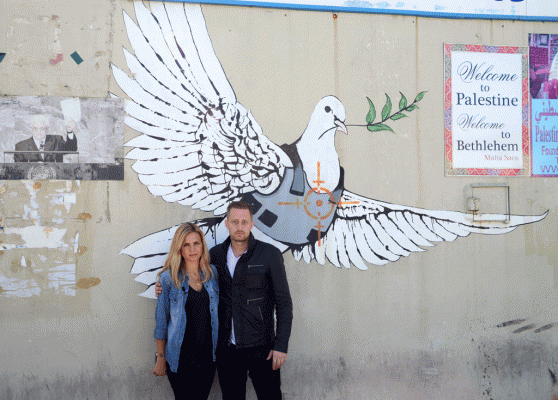 Foto e marrë nga gazetarja e Travel Channel Mariana Van Zeller, majtas, dhe prezantuesi Michael Voltaggio, i emisionit “Breaking Borders,” ku të dy kanë pozuar para një vepre arti që paraqet një pëllumb të pajisur me jelek antiplumb. Vepra është krijuar nga artisti i grafiteve Bansky në Betlehem, Palestinë. (AP Photo/Travel Channel)